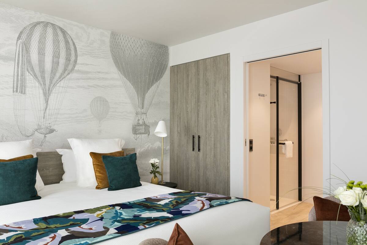 A modern hotel room with a large bed, a colorful abstract rug, a wooden wardrobe, and a glass shower door.
