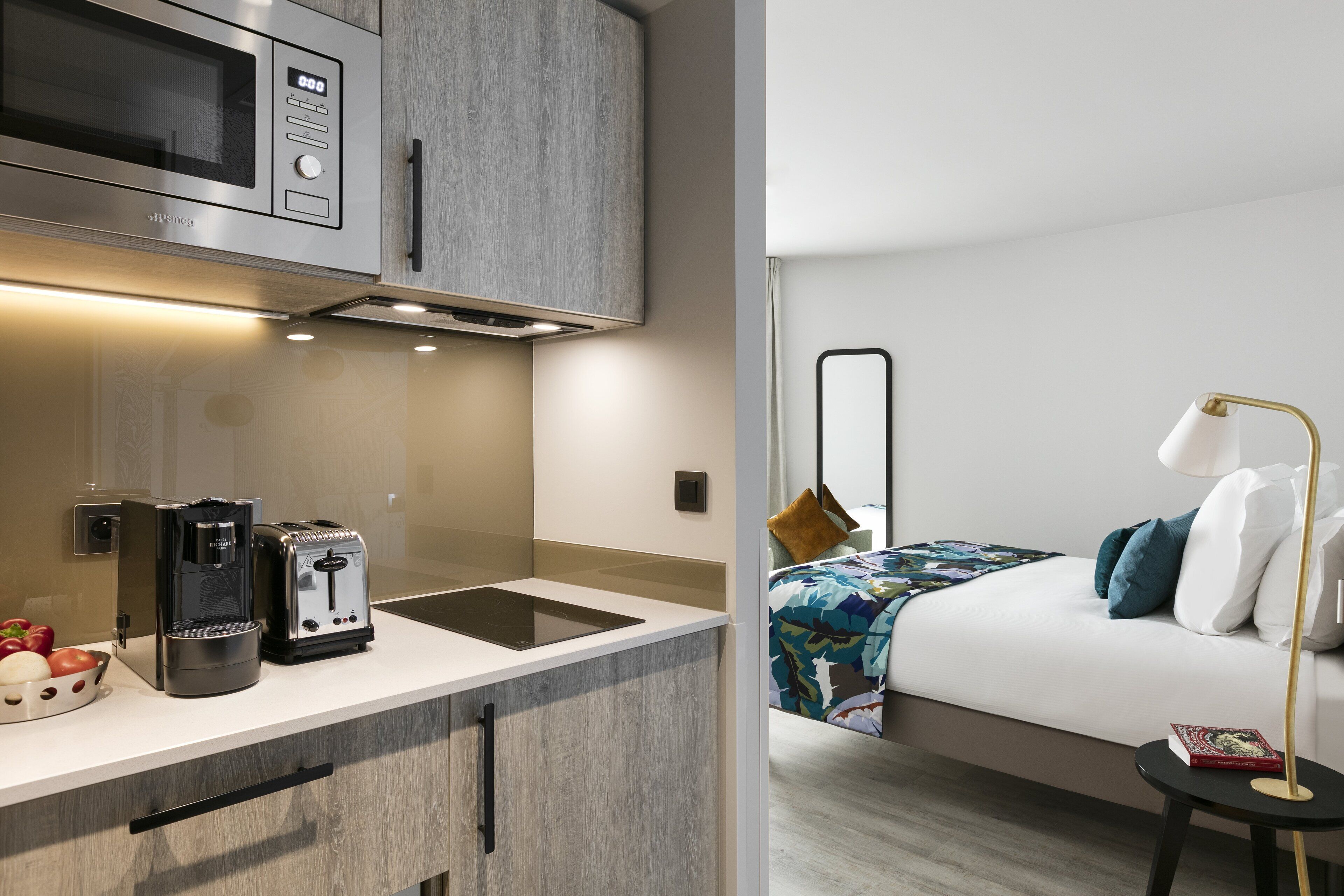A modern kitchen with a microwave, toaster, and coffee maker, and a bedroom with a bed, pillows, and a bedside lamp.