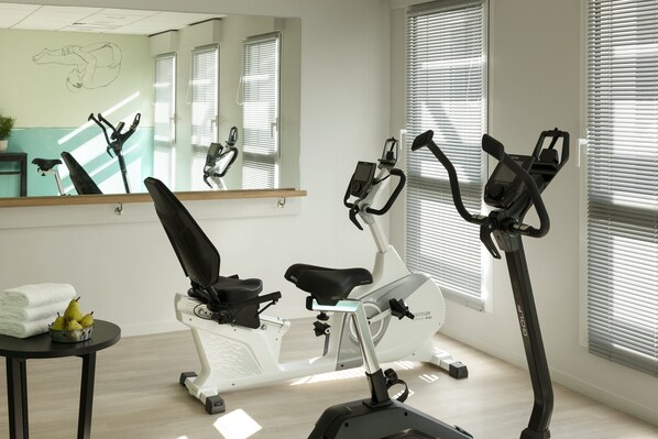 Un gimnasio con bicicletas estáticas, un espejo, una pequeña mesa con toallas y fruta, y ventanas con persianas.