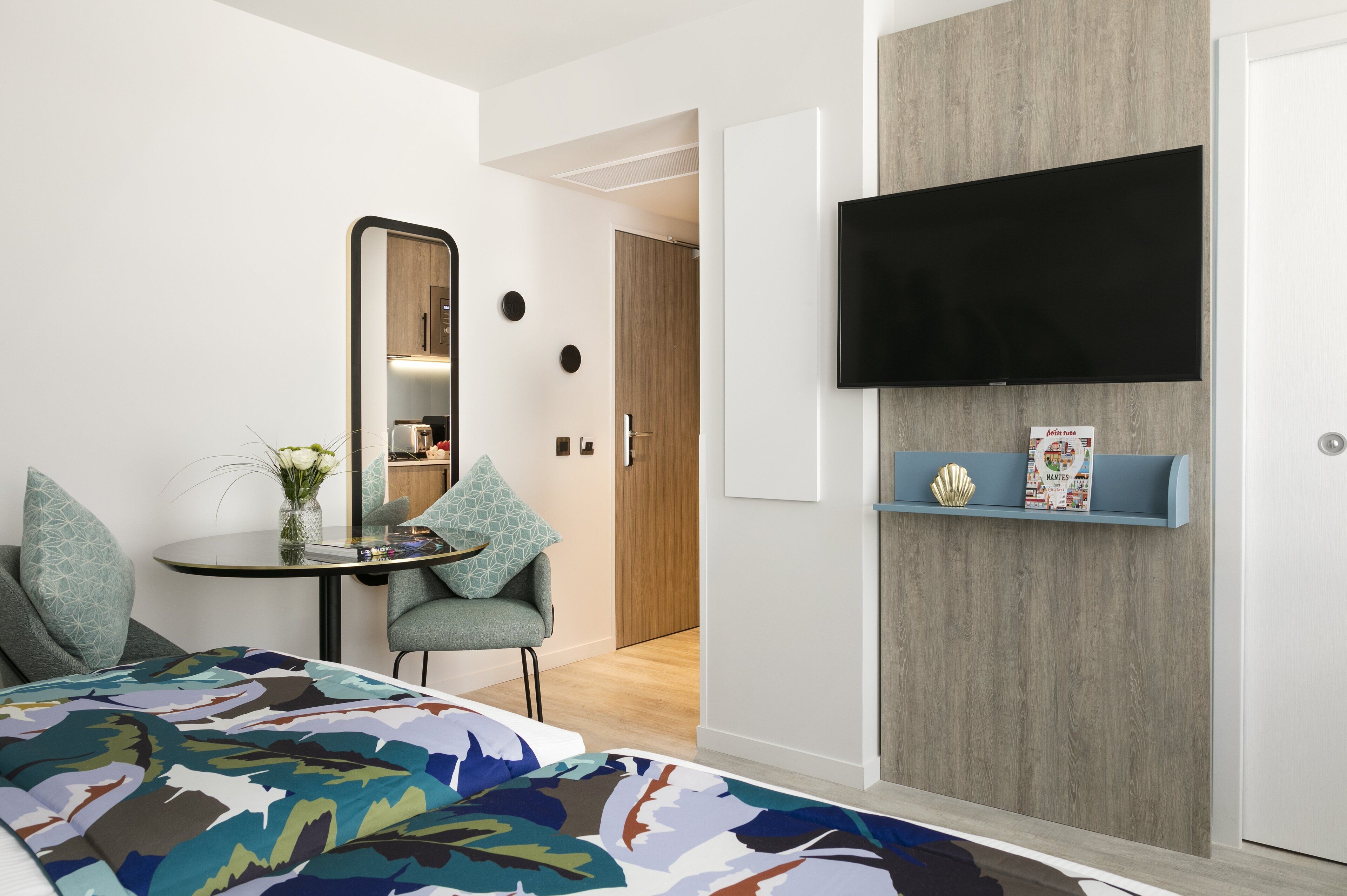 A modern hotel room with a bed, a small table with a vase, a chair, a TV mounted on the wall, and a wooden door.