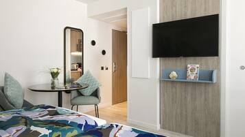 A modern hotel room with a bed, a small table with a vase, a chair, a TV mounted on the wall, and a wooden door.