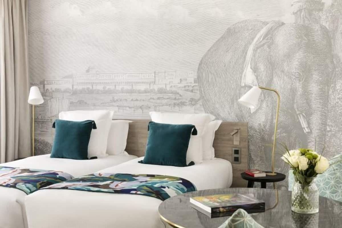 A hotel room with a bed, two bedside lamps, a round table with a vase of flowers, and a mural on the wall.