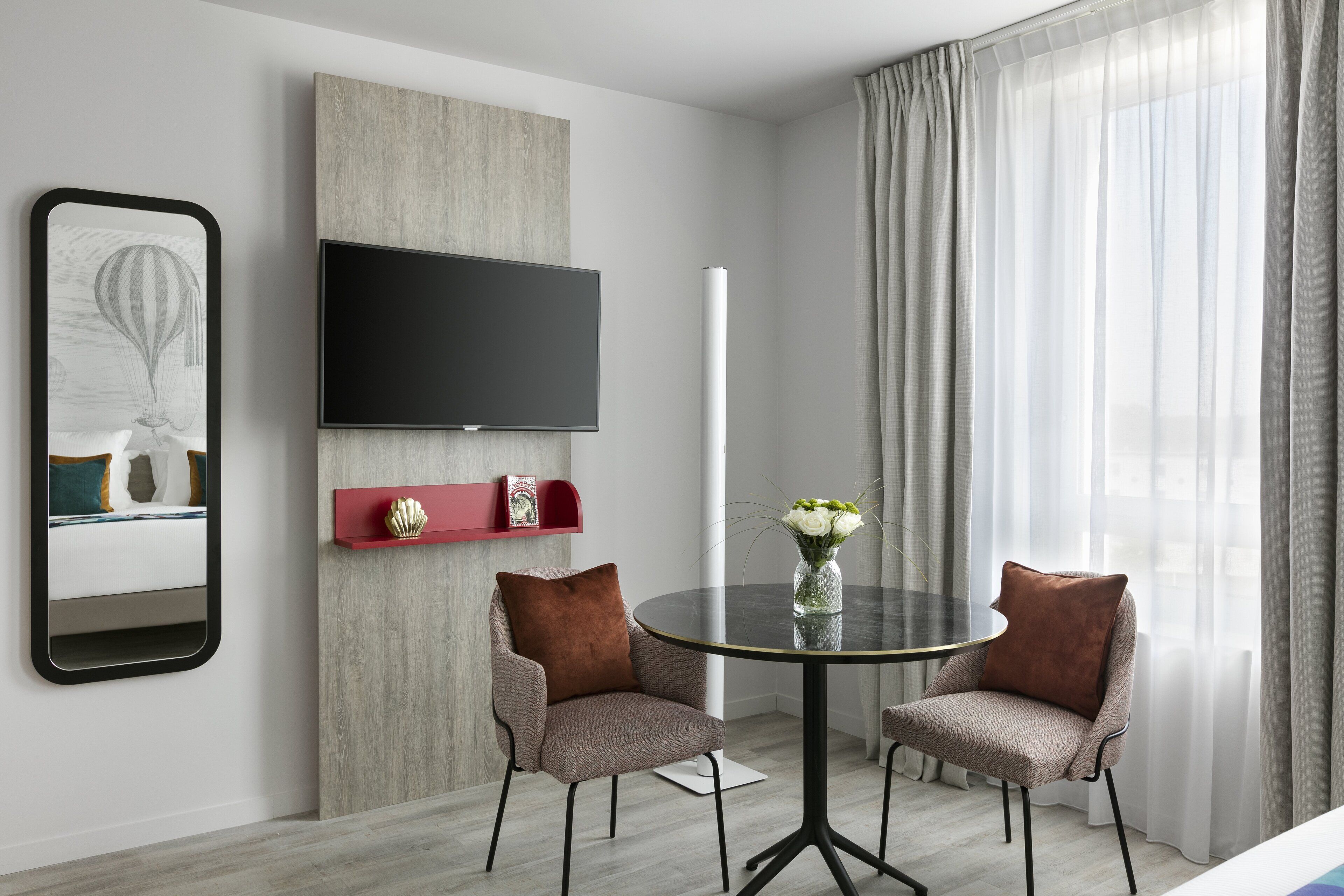 A modern hotel room with a flat-screen TV mounted on the wall, a round table with chairs, and a large window with curtains.