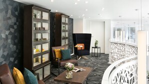 A modern hotel lobby with a geometric-patterned floor, a dark wood bookshelf, leather seating, and a black armchair. - Citadines Confluent Nantes (Nantes)