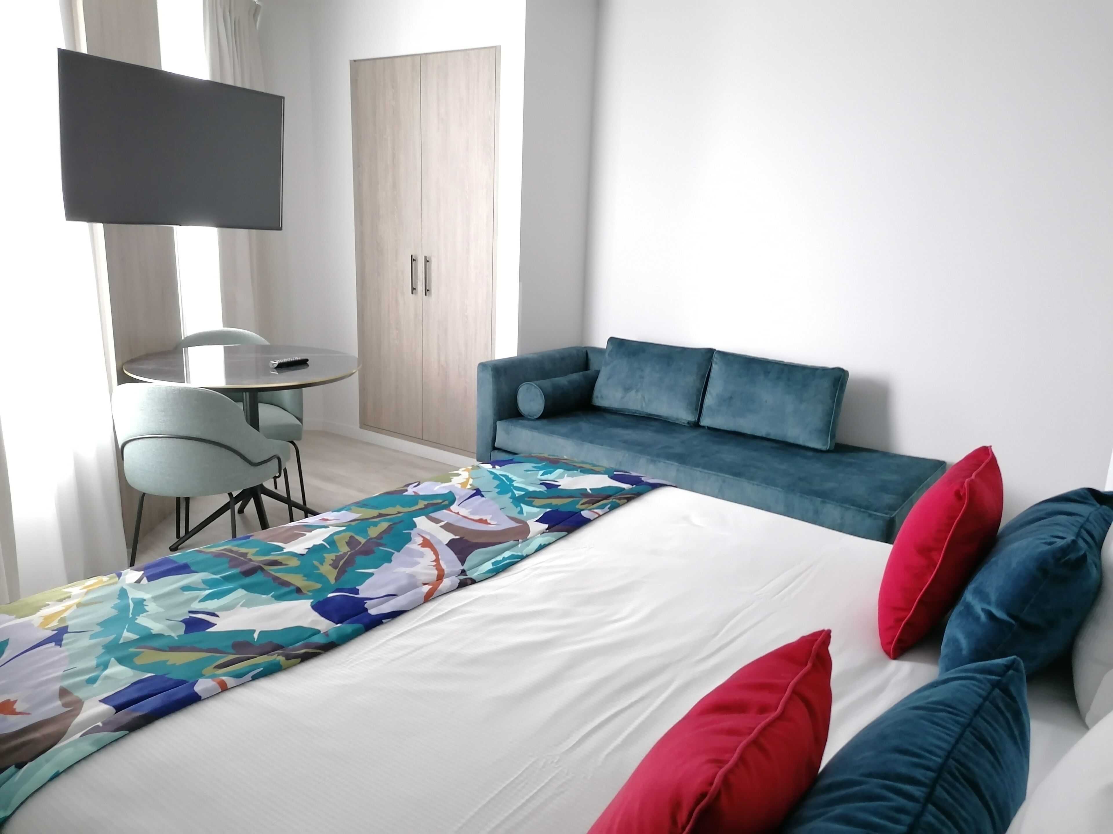 A modern hotel room with a bed, a blue sofa, a small round table, and a TV mounted on the wall.