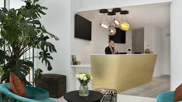 Uma área de recepção com recepcionista, uma grande planta, uma mesa com laptop e um lustre moderno.