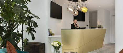 A reception area with a receptionist, a large plant, a desk with a laptop, and a modern chandelier.