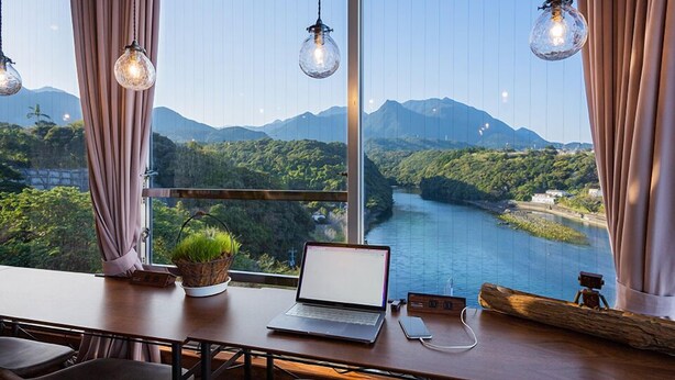 Ein Zimmer mit einem Laptop auf einem Schreibtisch, einer Pflanze und Blick auf einen Fluss und Berge.