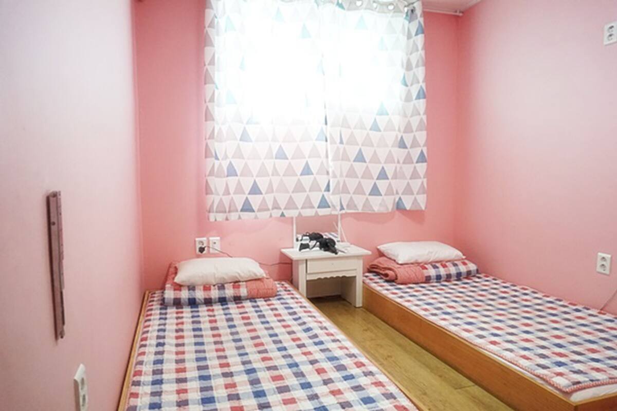 Un dormitorio pequeño y rosa con dos camas individuales, una mesita de noche y una ventana con cortinas estampadas.