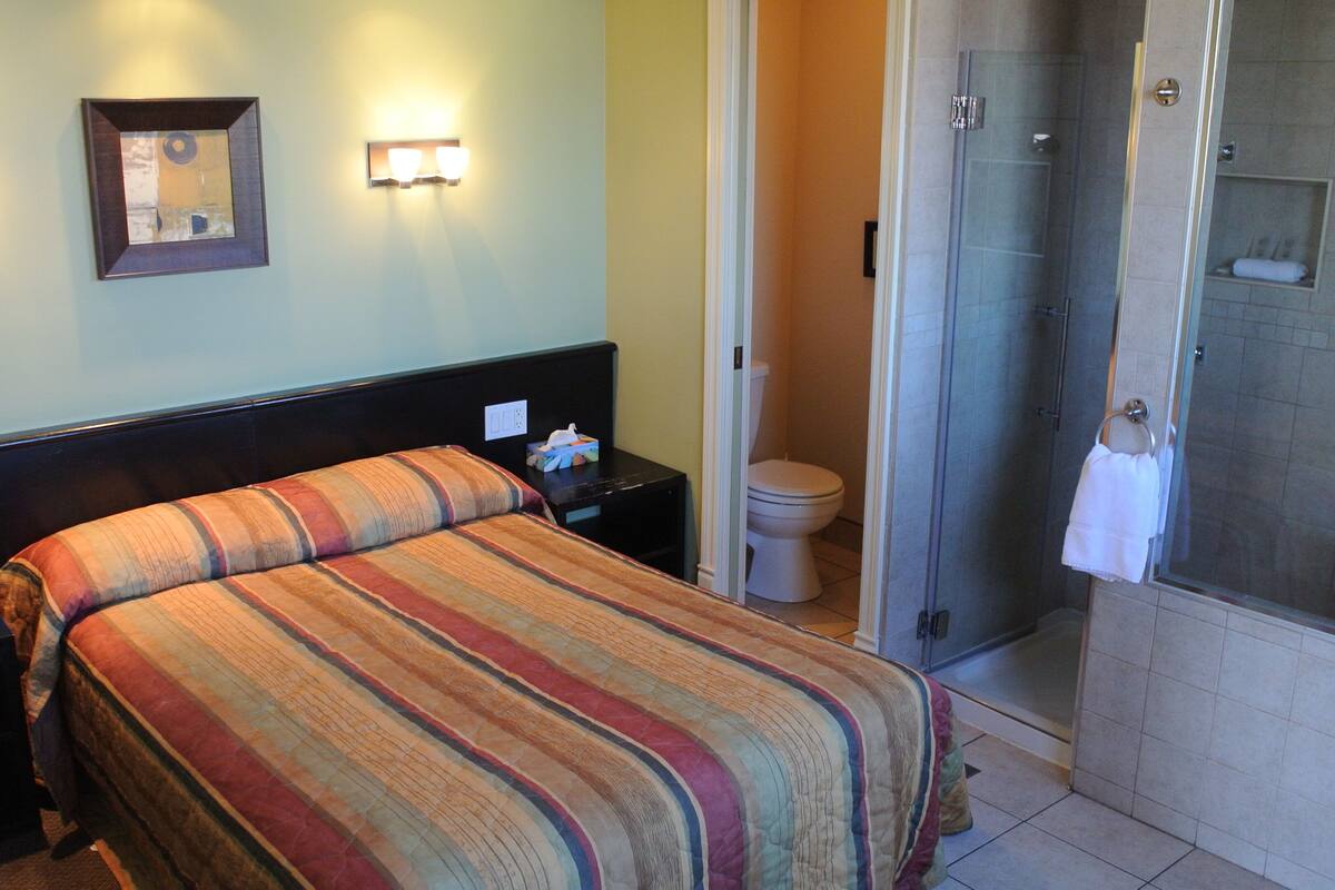 A hotel room with a bed, bedside table, wall-mounted lights, a framed picture, a toilet, and a shower area.