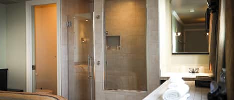 Un baño de hotel con ducha envuelta en vidrio, un lavamanos con artículos de aseo y una cama con manta a rayas.