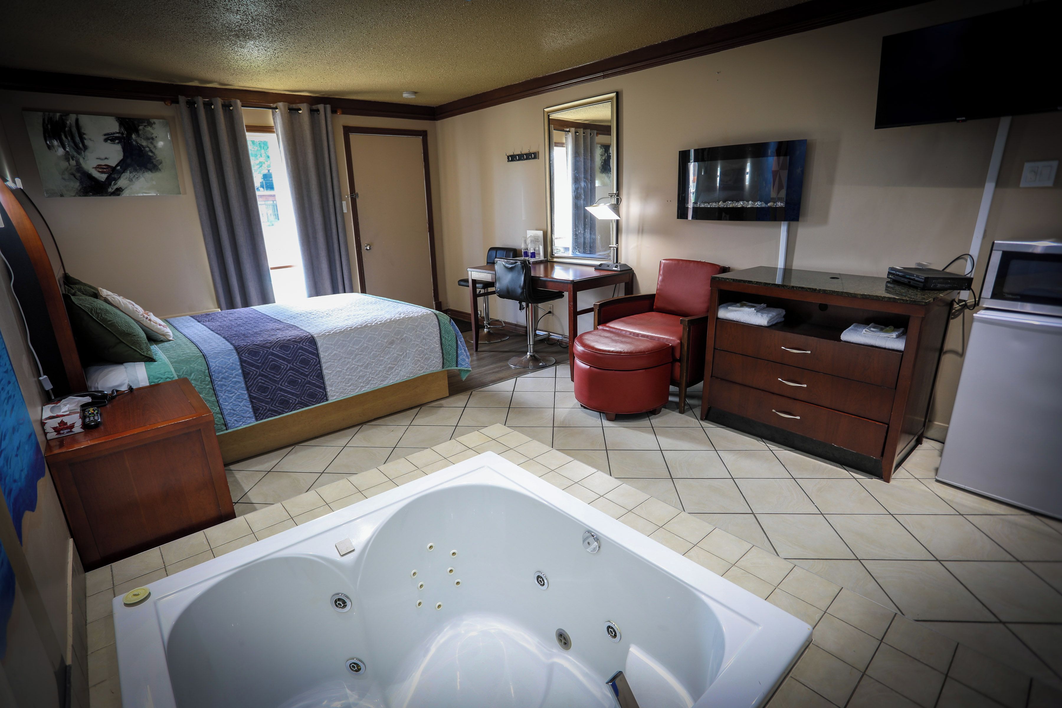A hotel room with a bed, a desk, a chair, a red leather chair, a television, a refrigerator, and a bathtub.