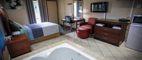 A hotel room with a bed, a desk, a chair, a red leather chair, a television, a refrigerator, and a bathtub.