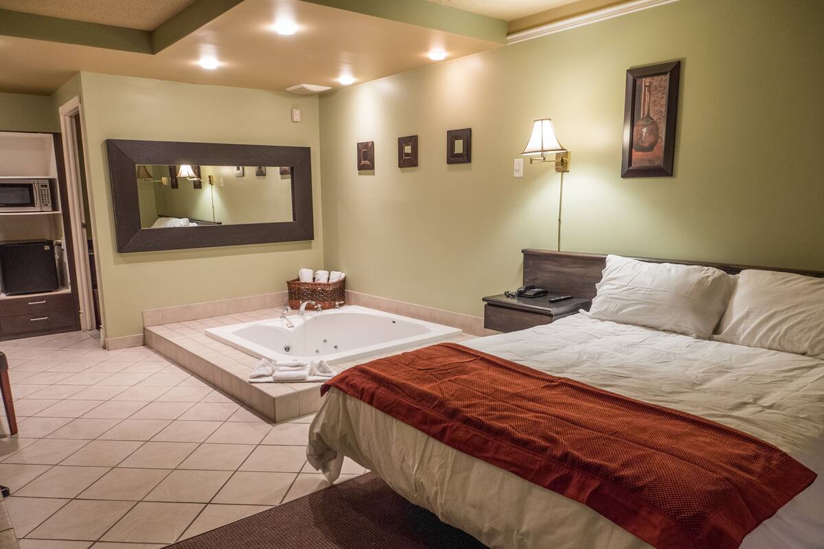 A hotel room with a large bed, a separate bathroom with a bathtub, a microwave, and a small table with a lamp.
