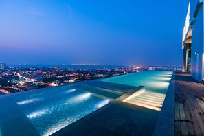 A rooftop infinity pool with a city view, a wooden deck, and a modern building facade. - YK Condotel Pattaya (Pattaya)