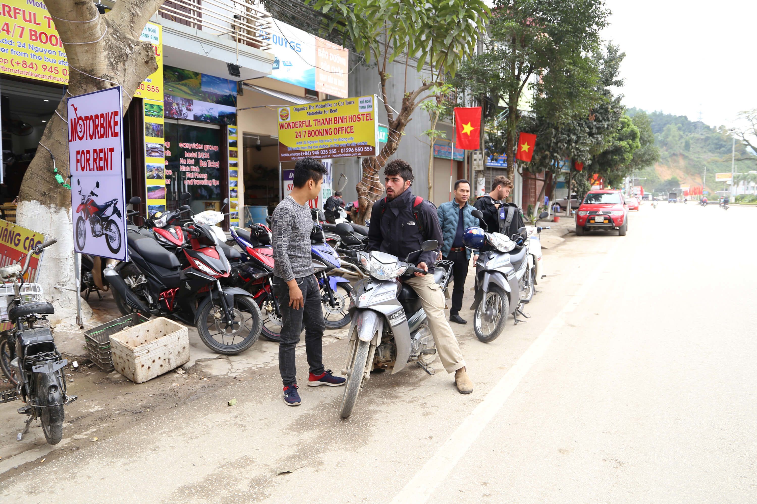 Una calle con motocicletas estacionadas al costado, un letrero que anuncia un servicio de alquiler de motos, y un albergue con una oficina de reservas abierta las 24 horas.
