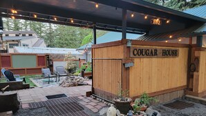 Un espace extérieur accueillant, aménagé avec des sièges, et une cabane en bois portant l’inscription « COUGAR HOUSE », le tout entouré de plantes en pot et bordé d’un chemin de gravier.