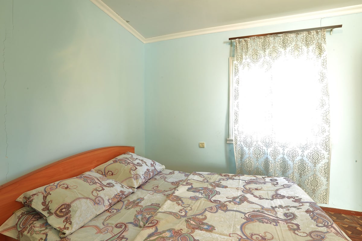 Ein Schlafzimmer mit einem Bett, einem hölzernen Kopfteil, einer gemusterten Bettdecke und einem Fenster mit Vorhang.