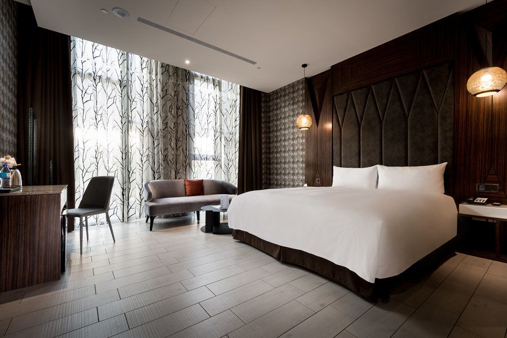 A modern hotel room with a large bed, a sofa, a desk, and a view through sheer curtains.