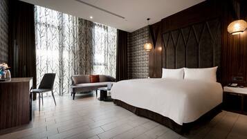 A modern hotel room with a large bed, a sofa, a desk, and a view through sheer curtains.