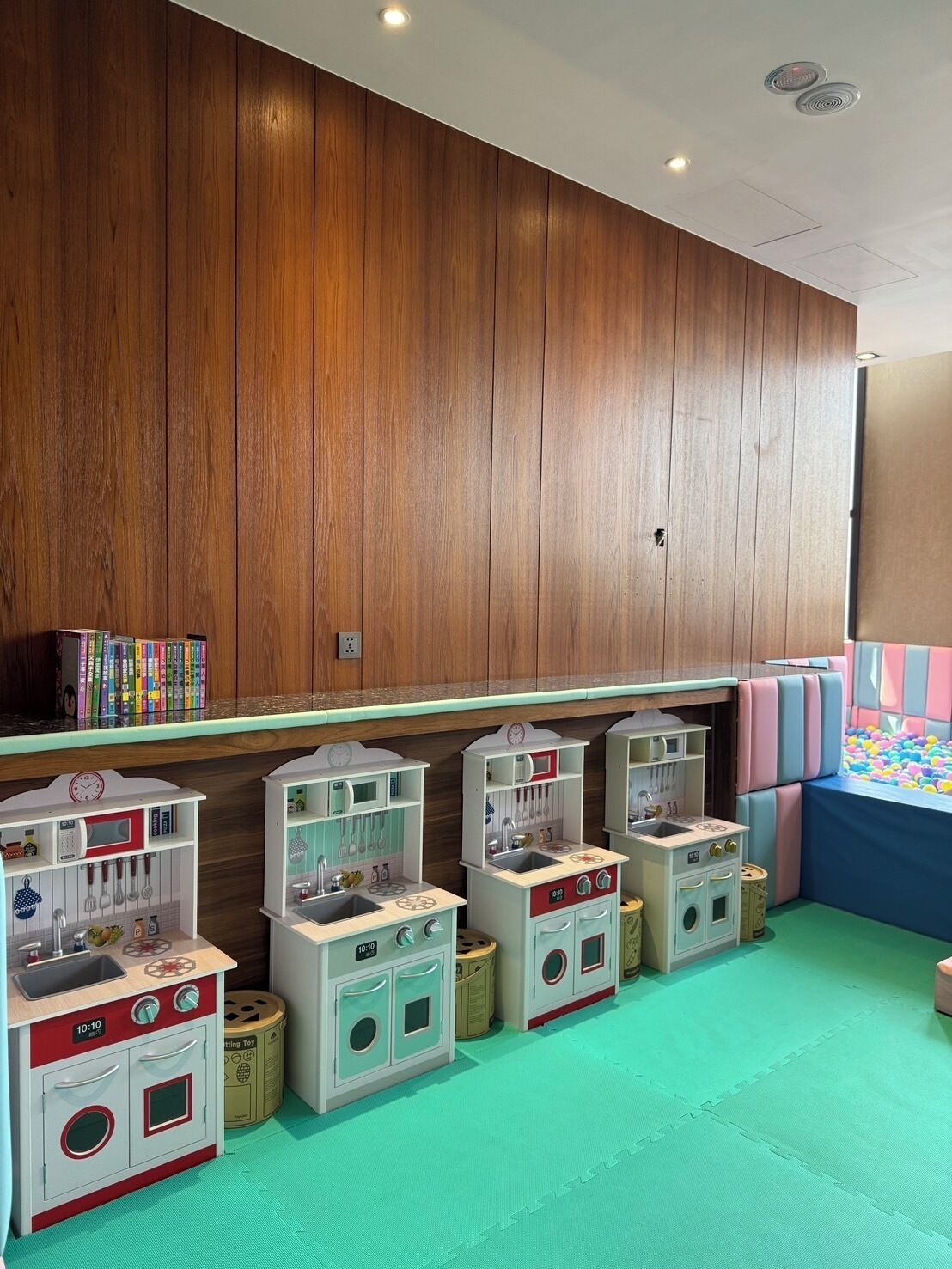 A children's play area with wooden panel walls, a green floor, and multiple play kitchens.