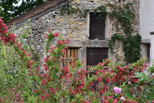 Um edifício rústico de pedra com porta e janela de madeira, rodeado por vegetação exuberante e plantas floridas.