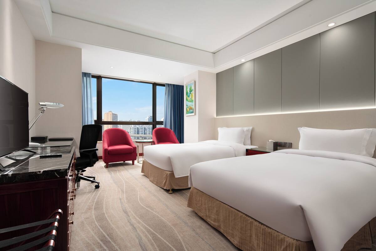 A hotel room with two beds, a desk, a TV, and a red chair.