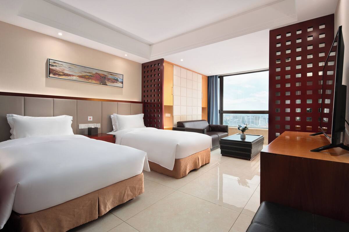 A modern hotel room with two beds, a desk, a chair, and a large window offering a city view.