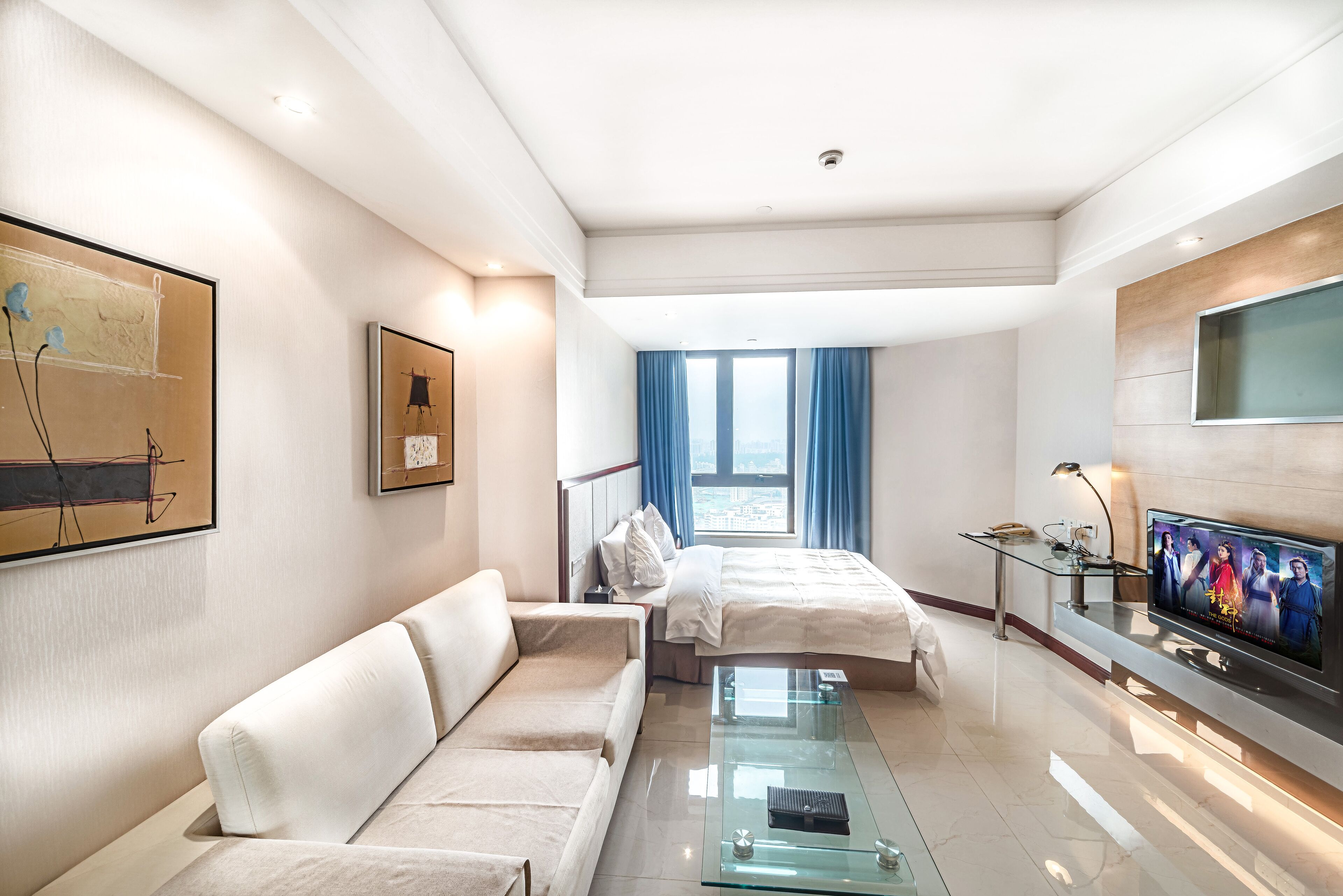 A modern hotel room with a glass coffee table, a sofa, a bed, a TV, and artwork on the walls.