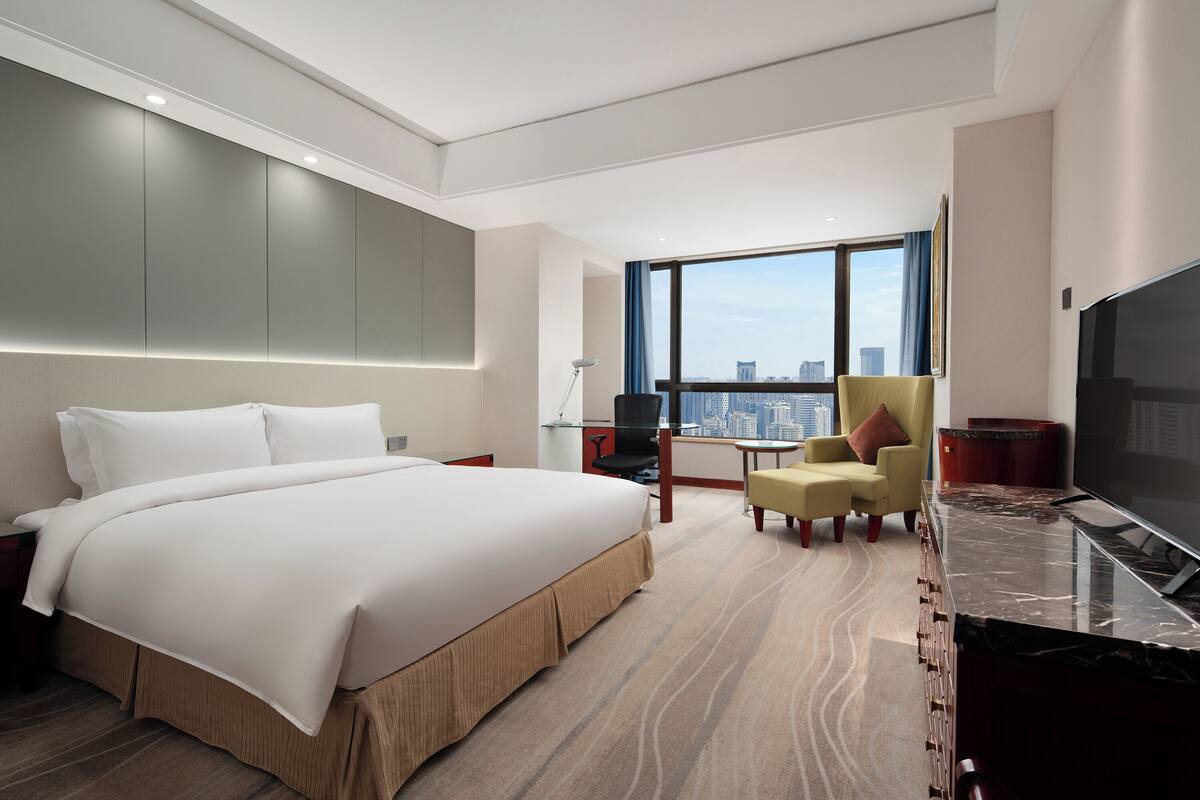 A hotel room with a large bed, a desk with a chair, a TV, and a view of the city.