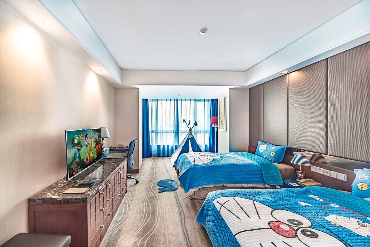 A hotel room with two beds, a TV, a desk, and a window with curtains.