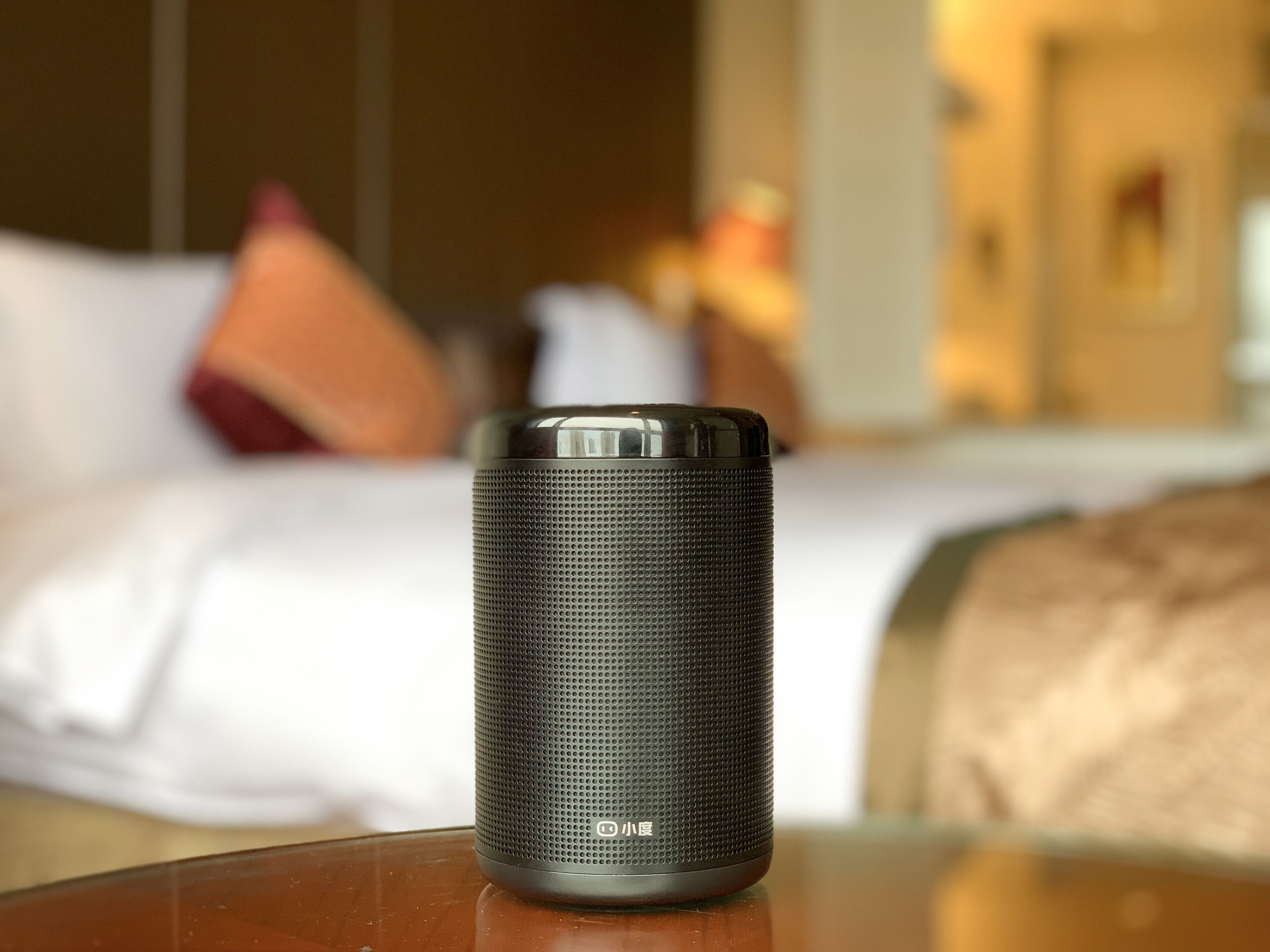 Ein zylindrischer Lautsprecher mit Struktur auf einer Holzfläche in einem Hotelzimmer, im Hintergrund ein Bett mit Kissen.