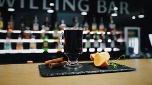 Een donker drankje in een glazen mok op een zwarte leisteen onderzetter, met kaneelstokjes en een sinaasappelschijfje op een houten tafel.