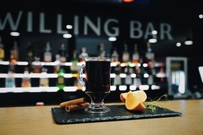 A dark beverage in a glass mug on a black slate coaster, accompanied by cinnamon sticks and an orange slice on a wooden table. - Willing Hotel (Minsk)