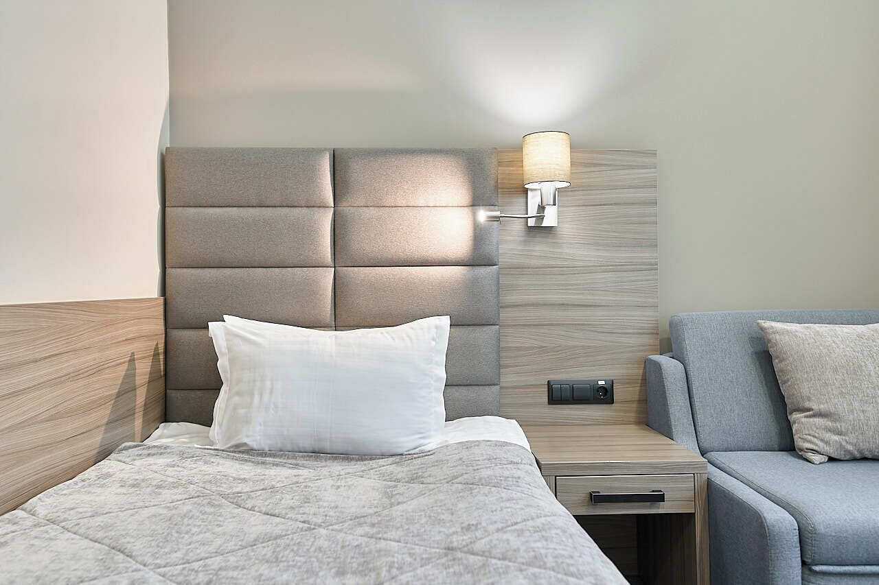 A modern hotel room with a tufted headboard, a bedside table with a lamp, a grey sofa, and a bed with a quilted cover.