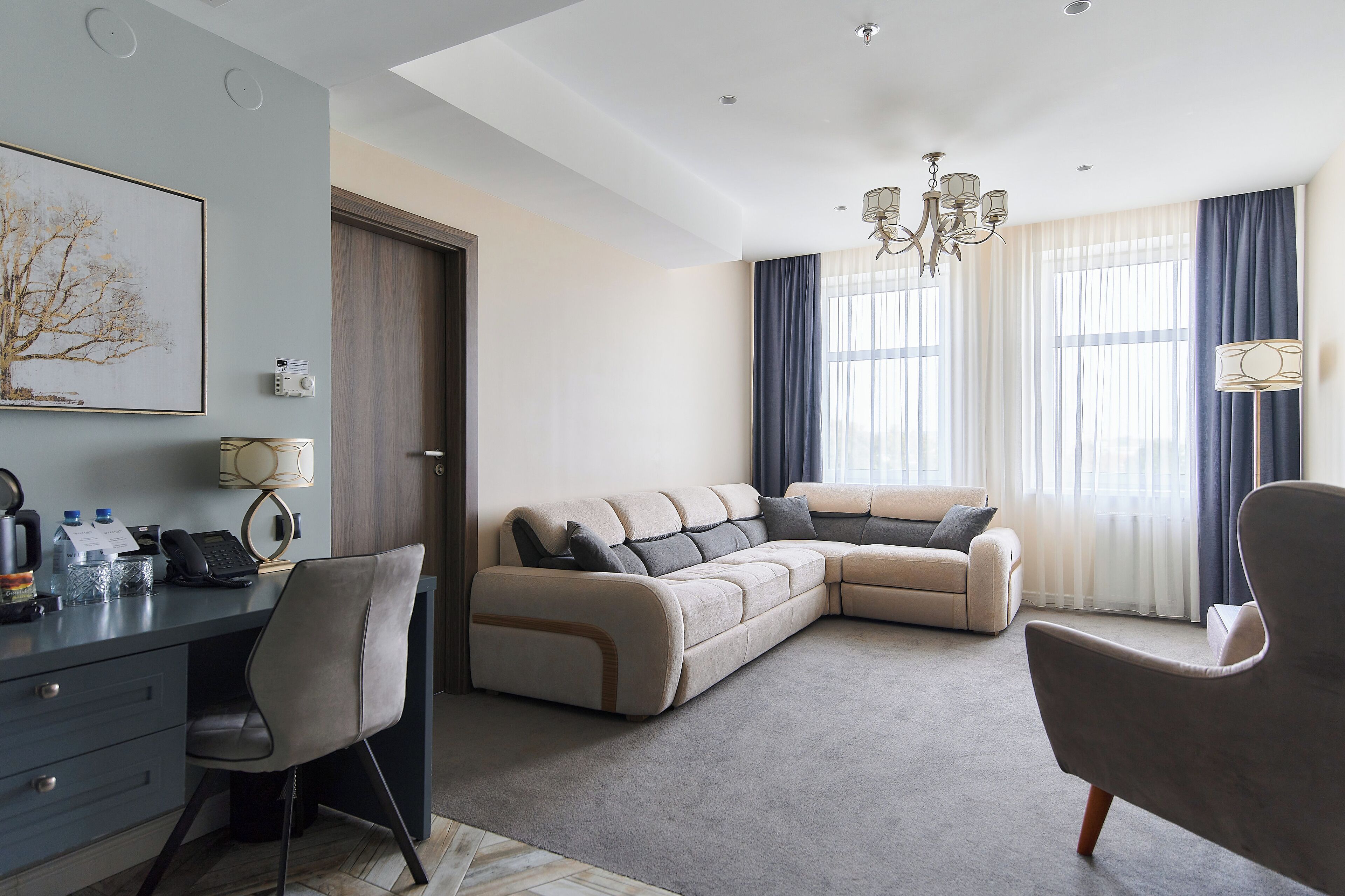 A modern living room with a sectional sofa, a desk with a chair, a chandelier, and large windows with curtains.