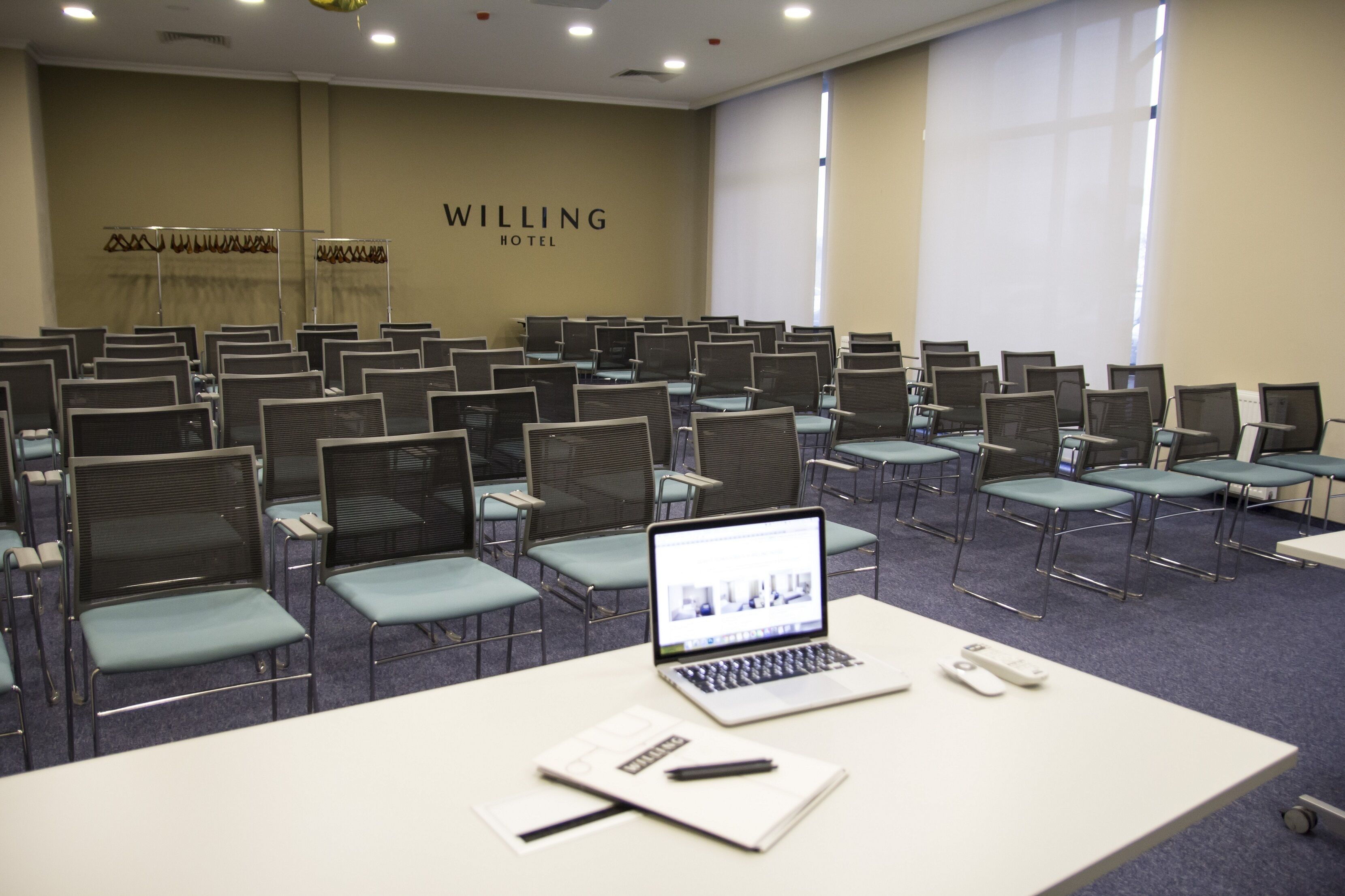 A conference room with rows of chairs, a laptop on a table, and a sign reading "WILLING HOTEL".