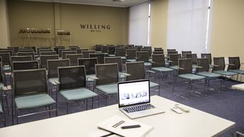A conference room with rows of chairs, a laptop on a table, and a sign reading "WILLING HOTEL".