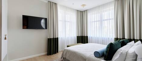 A bedroom with a bed, a flat-screen TV mounted on the wall, and a window with sheer curtains.