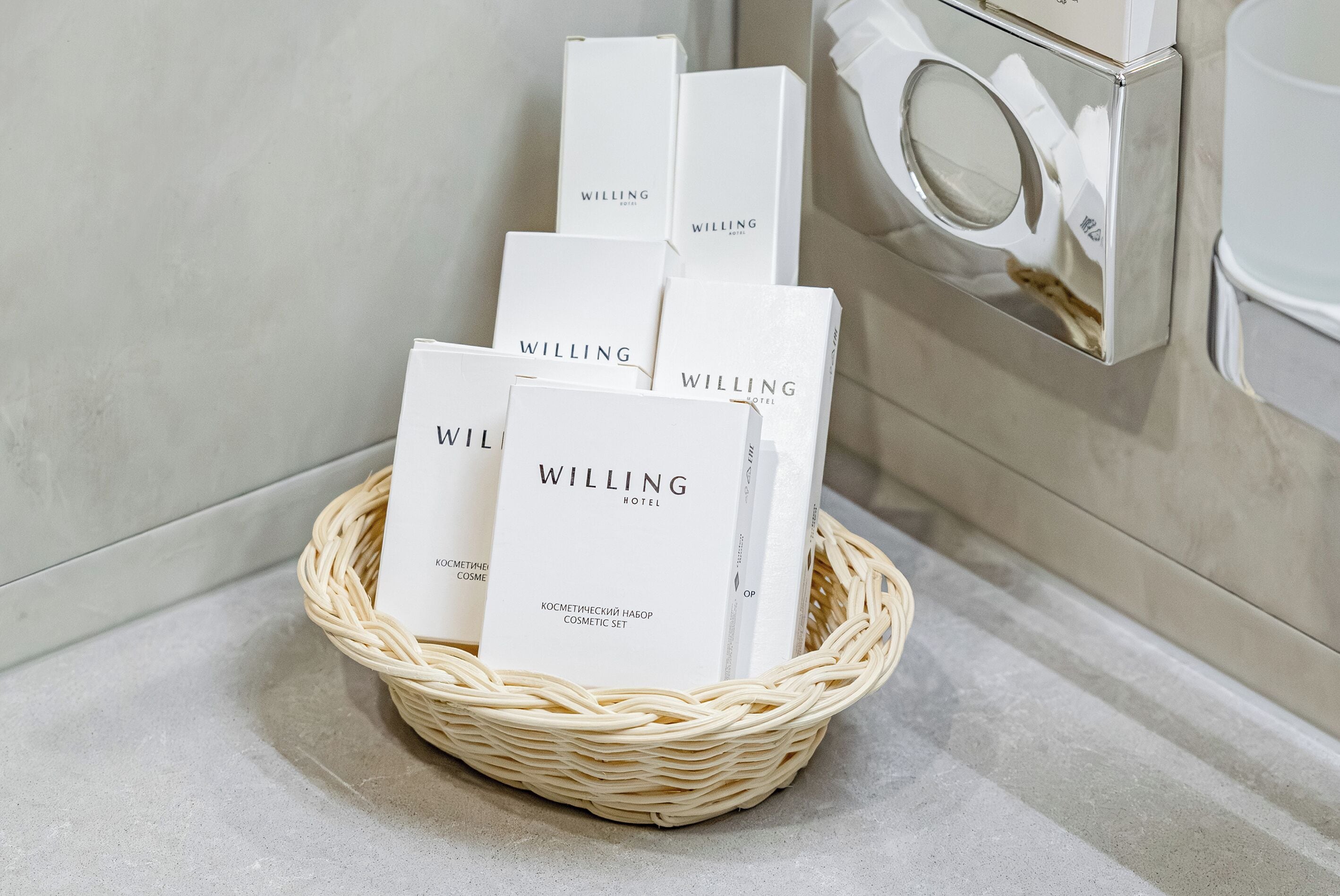 Una cesta de mimbre con varias cajas blancas etiquetadas como “WILLING HOTEL” sobre una encimera de baño.
