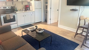 A modern kitchen with white cabinets, a built-in oven, and a microwave. There is a dining area with a dark coffee table and a swan-shaped decorative item. A television is mounted on the wall, and a chair is visible in the corner. - 1 bed/1 bath, 1.5 blocks to beach/town, dog friendly (Union Pier)