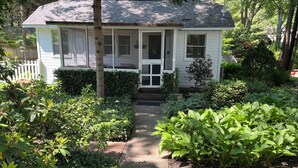 A small white house with a porch, surrounded by lush greenery and a well-maintained garden.