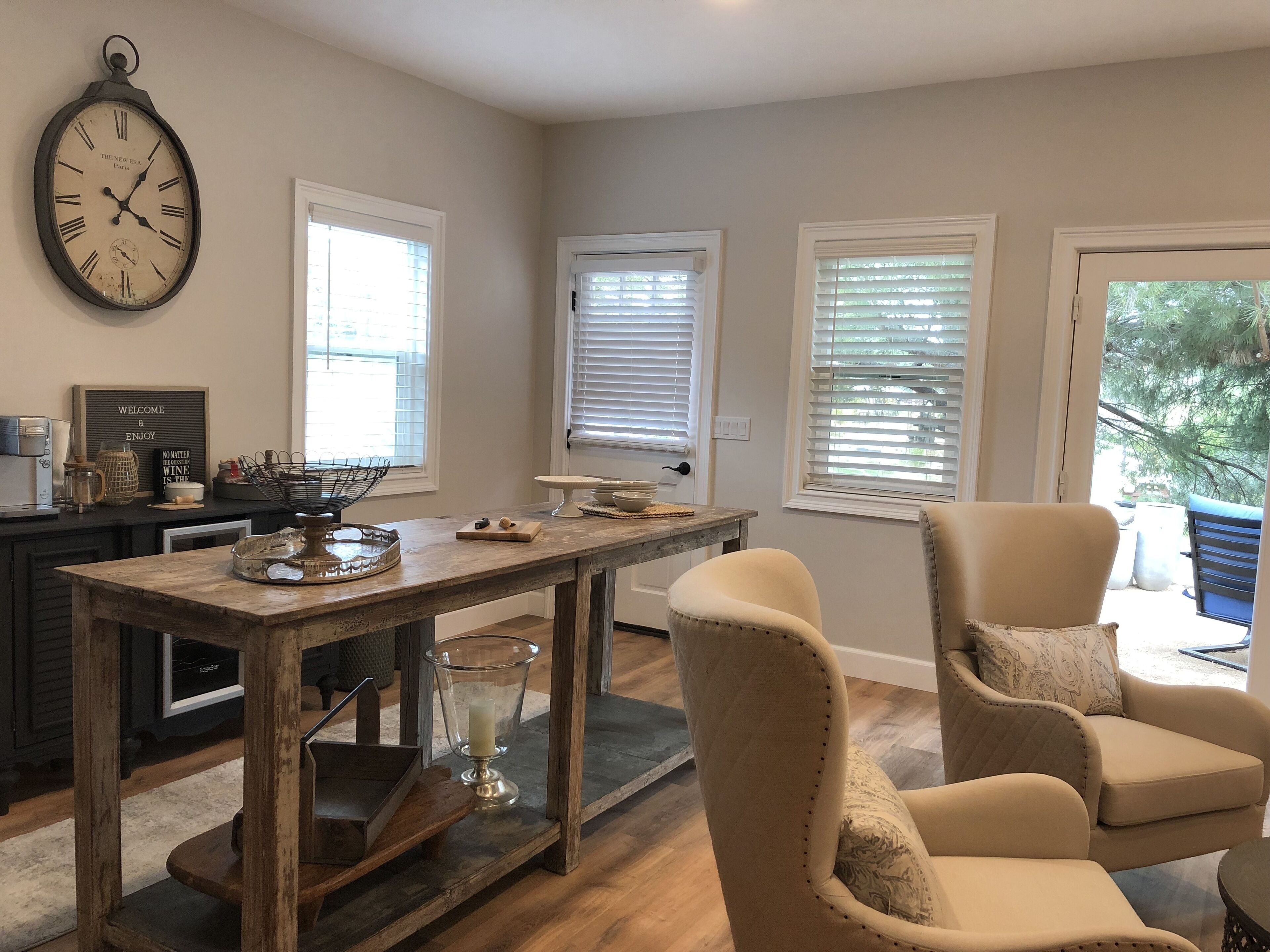 A dining area with a wooden table, two beige armchairs, a clock on the wall, and windows with blinds.