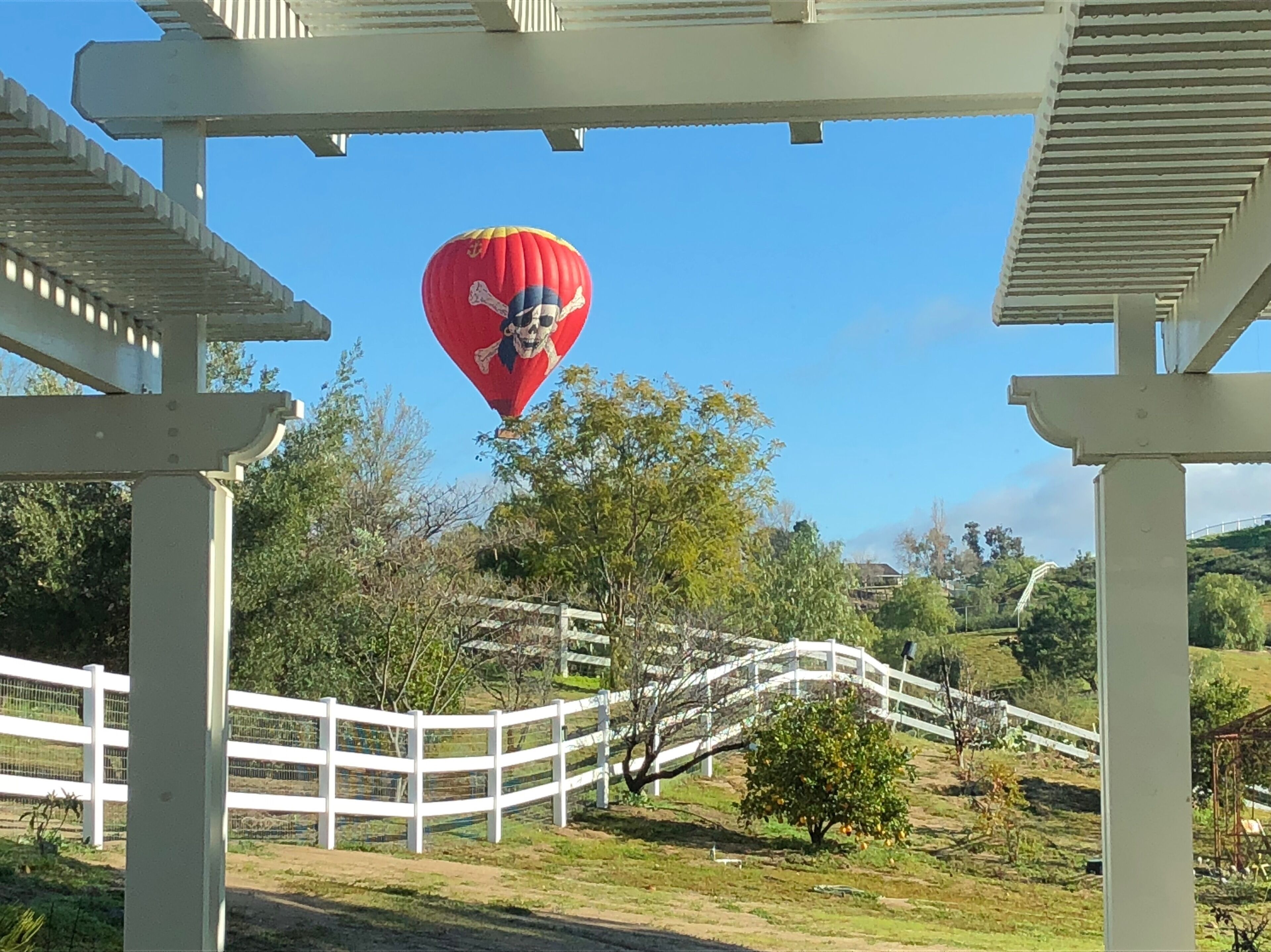 A hot air balloon with a skull design flying above a white wooden bridge in a rural setting.