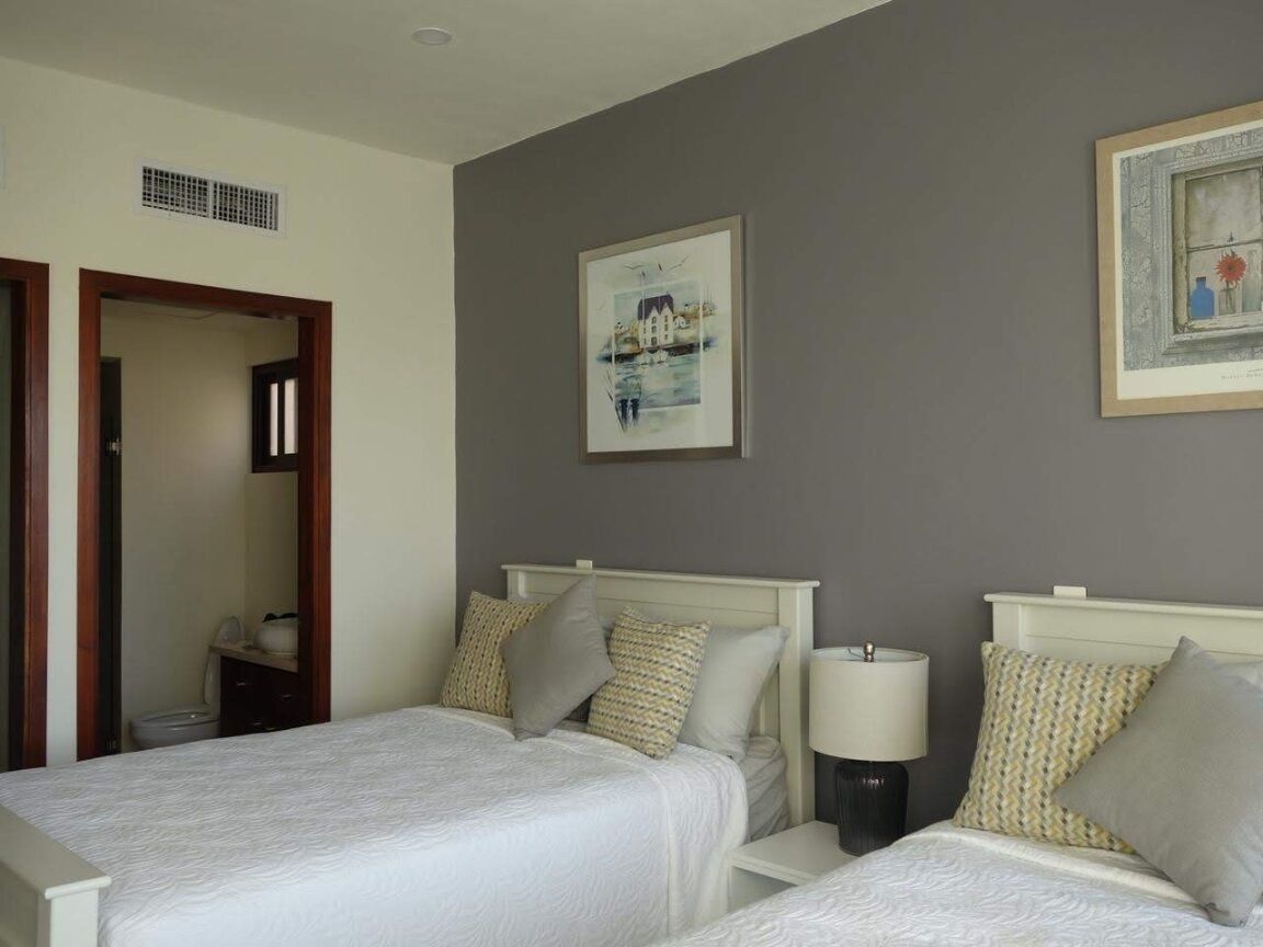 Habitación de hotel con dos camas, una mesita de noche con una lámpara, un cuadro enmarcado en la pared y una puerta que da a otra habitación.