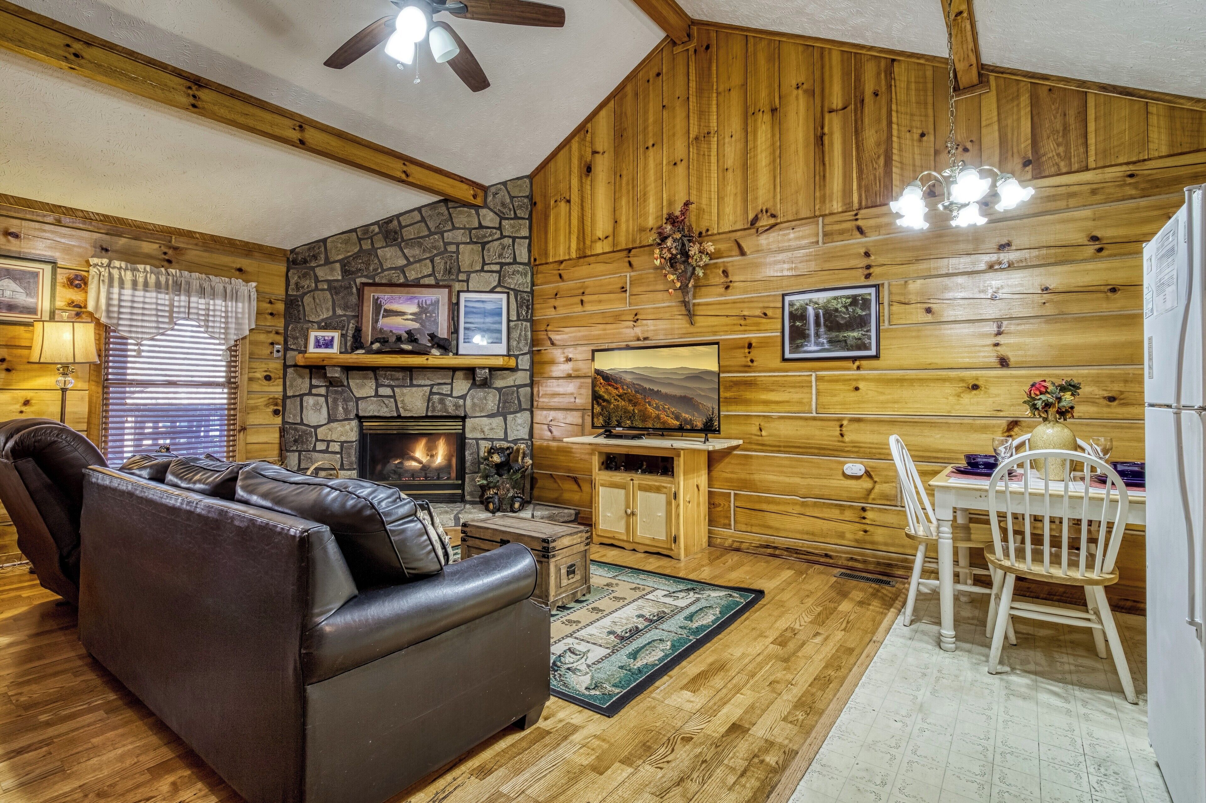 A cozy cabin interior with a stone fireplace, wooden walls, a leather sofa, and a dining area with a white table and chairs.
