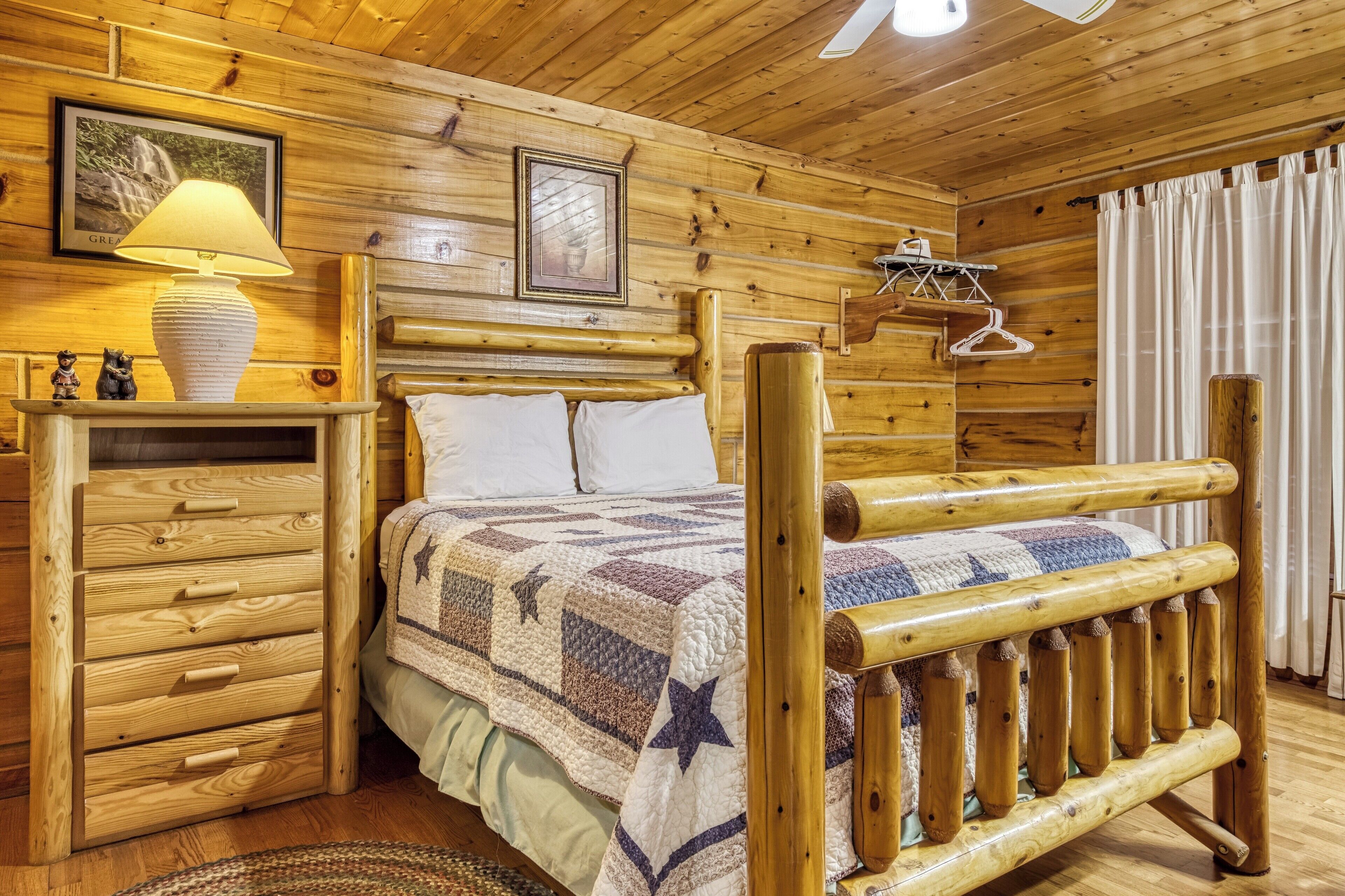 A wooden cabin room with a bed, a dresser, a lamp, and a framed picture on the wall.