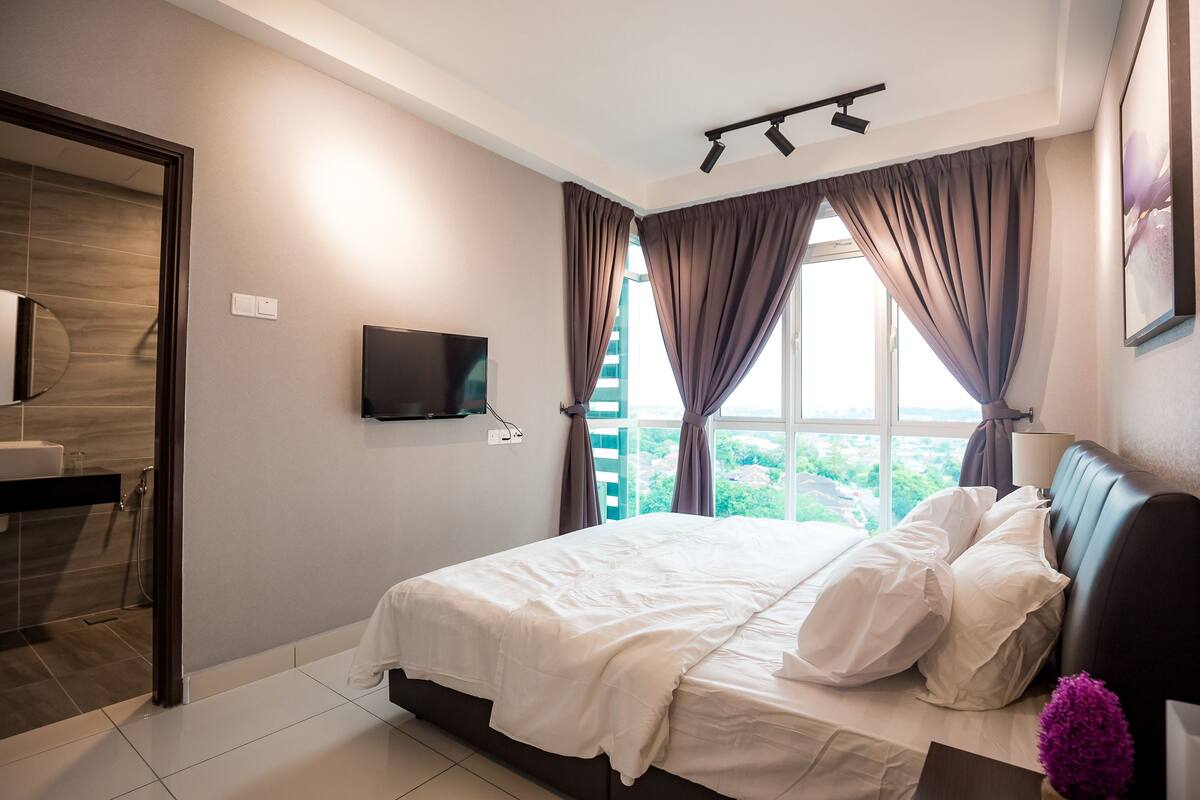 Ein Schlafzimmer mit Bett, Fernseher, einem Fenster mit Vorhängen und Blick auf Grünflächen.