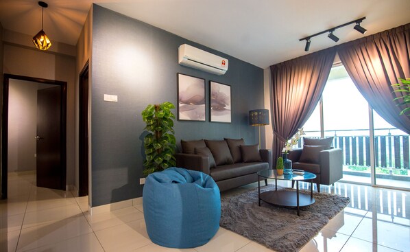 A modern living room with a sofa, a blue bean bag, a coffee table, and a large window with curtains. - Taman Daya Homybed Homestay (Johor Bahru)