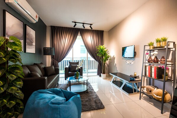 A modern living room with a sofa, coffee table, and a blue bean bag. There is a TV mounted on the wall, a bookshelf, and a potted plant. - Taman Daya Homybed Homestay (Johor Bahru)